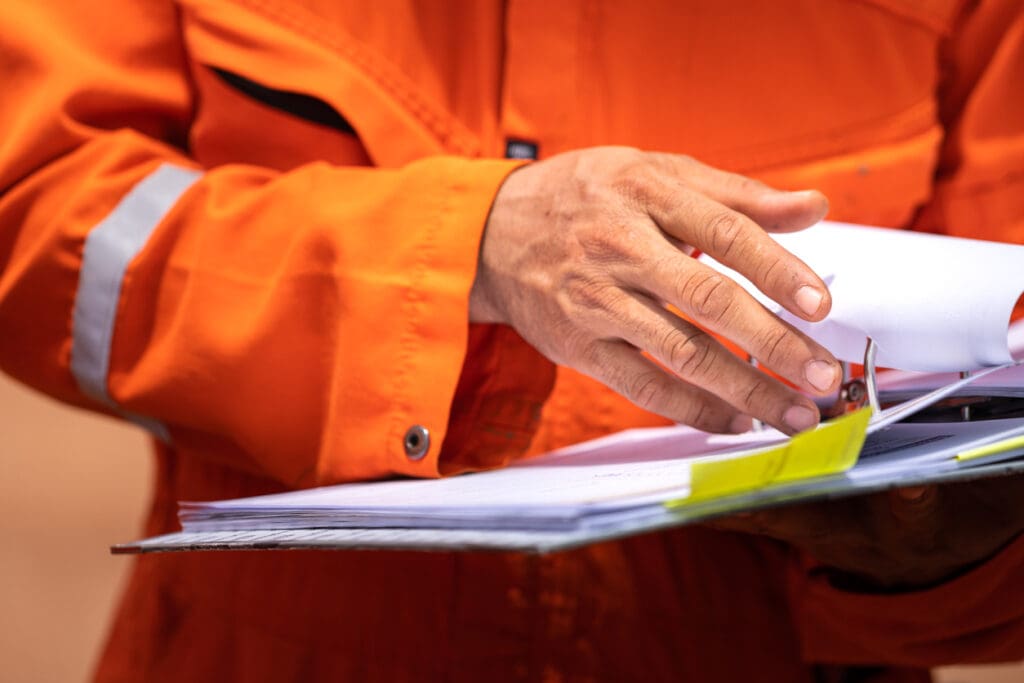 A worker in orange coverall uniform is opening documents to make a discussion on working procedure, safety audit in operation action photo.